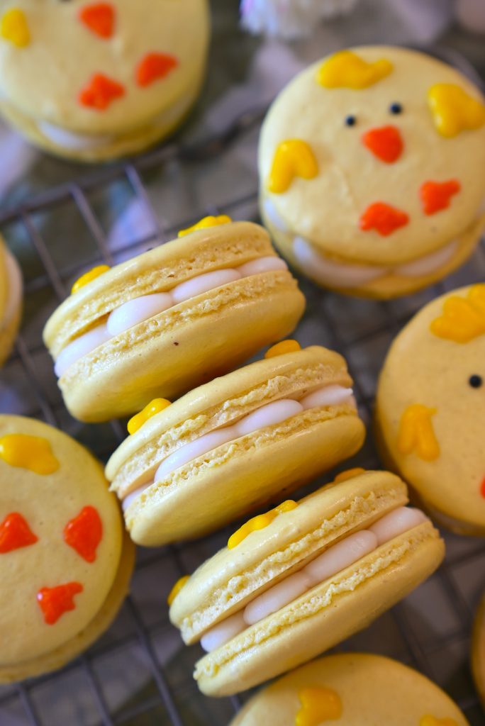 Yellow macarons with white filling are decorated with orange and yellow icing to resemble chick faces, displayed on a wire rack.
