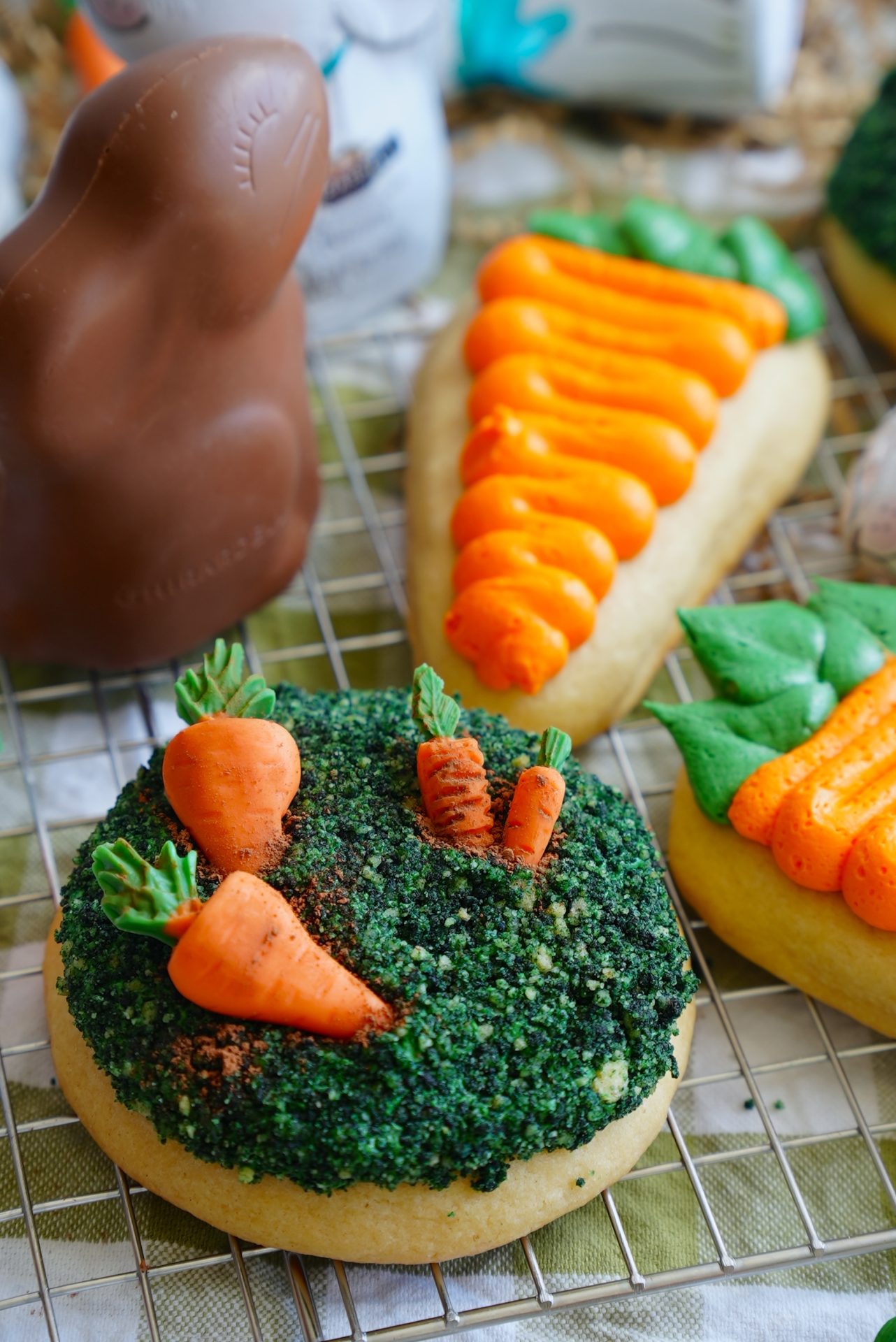 A round cookie topped with green sprinkles and decorated with small icing carrots sits on a cooling rack next to a carrot-shaped cookie and a chocolate bunny.