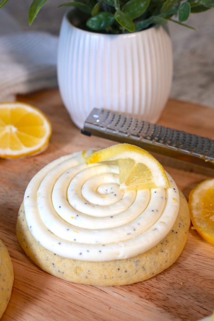 A lemon poppy seed cookie topped with swirled white icing and a lemon slice, placed on a wooden board with lemon slices, a grater, and a potted plant in the background.