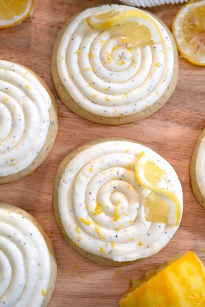 Round cookies with white swirled frosting, poppy seeds, and garnished with lemon zest and lemon slices, displayed on a wooden surface.