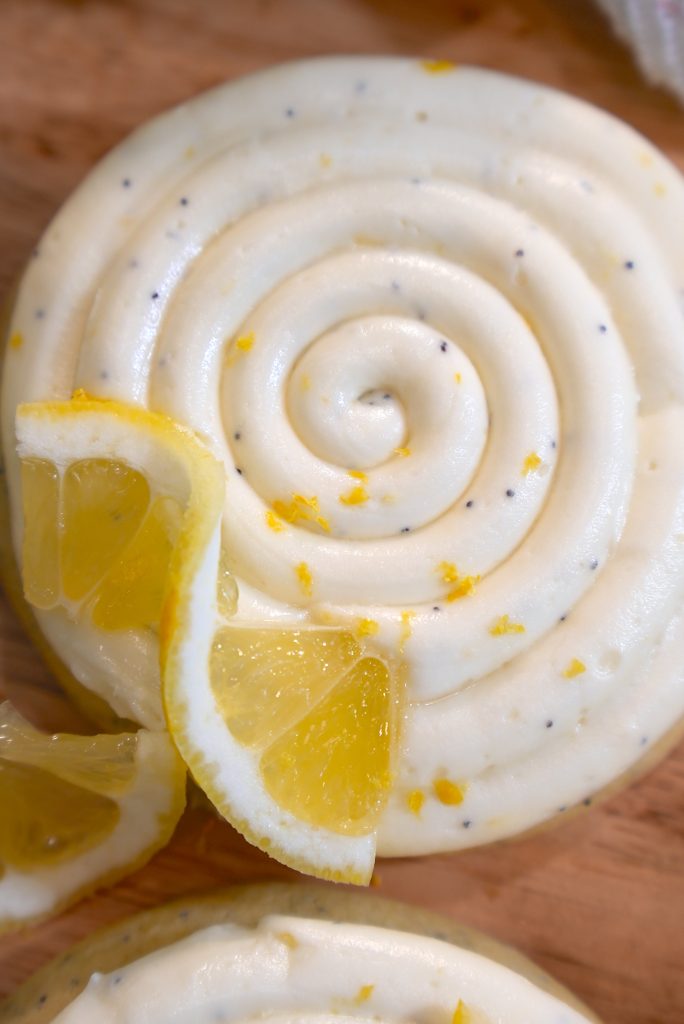 A close-up of a swirled lemon poppy seed frosting topped with lemon zest and two lemon slices, displayed on a wooden surface.