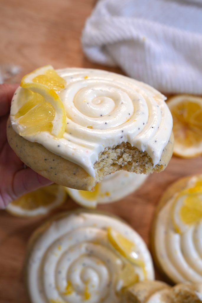 A hand holds a lemon poppy seed cookie topped with swirled white icing and lemon slices. A bite is taken out of the cookie. More similarly decorated cookies are on the wooden surface below.