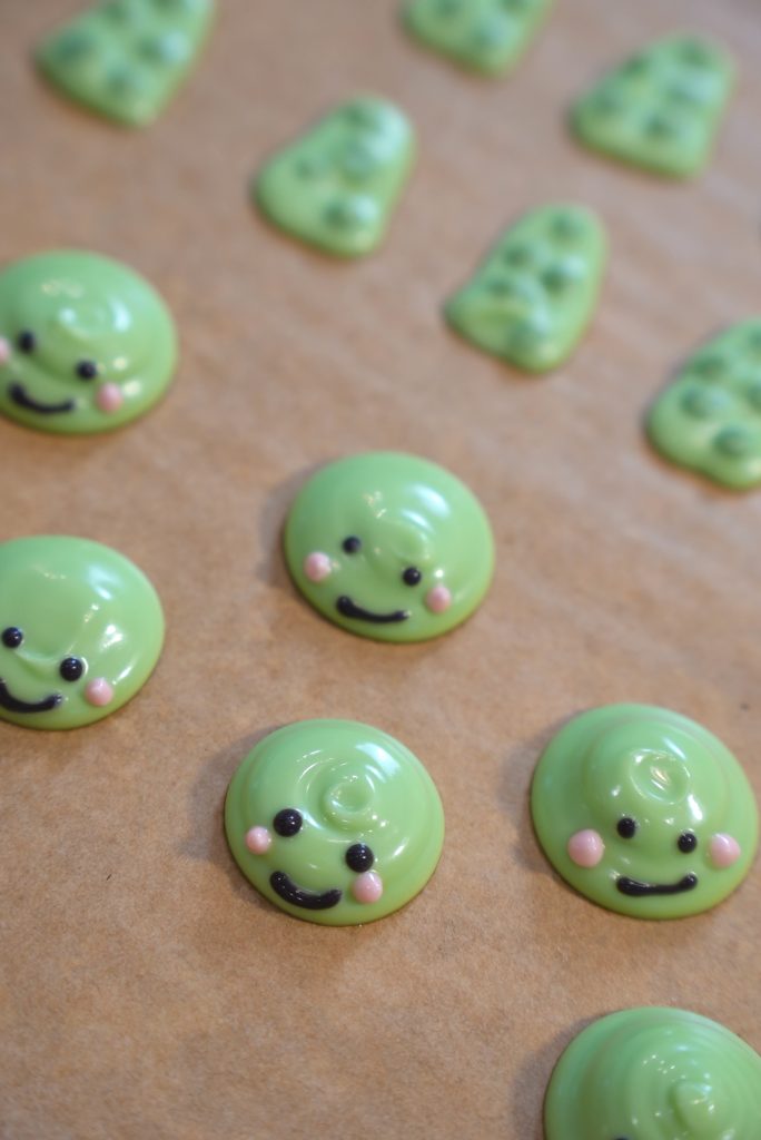 Green candy blobs with smiling faces and pink cheeks are arranged on a sheet of brown parchment paper. Some candies have a swirled top, and others are shaped like trees with decorated tops.