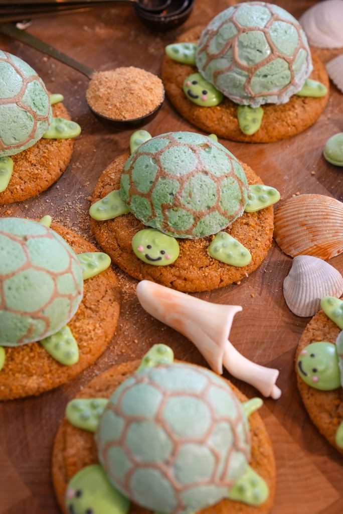 Turtle-themed cookies with green turtle-shaped decorations on top, arranged on a wooden surface with sea shells and a spoonful of brown sugar nearby.