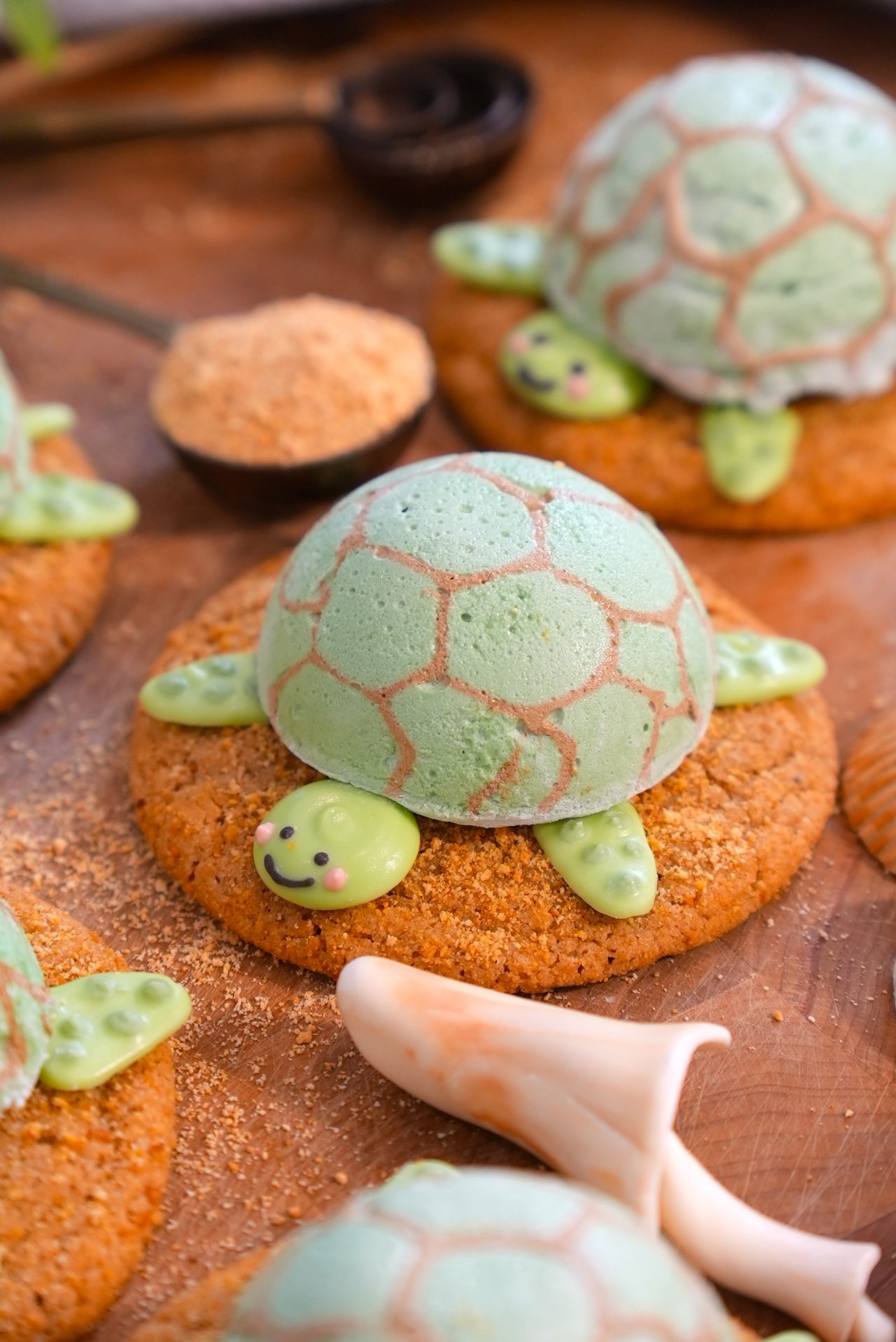 Adorable turtle-shaped cookies with green shells and smiling faces are placed on round cookies, resembling turtles sitting on sand. A spoon with brown sugar and cookie crumbs are also visible in the background.