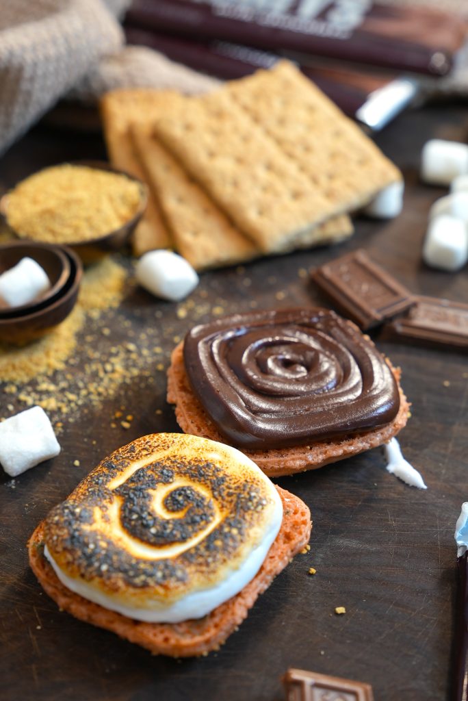 Two s’mores with toasted marshmallows and swirled chocolate on graham crackers are on a wooden surface, surrounded by marshmallows, graham crackers, and chocolate pieces.