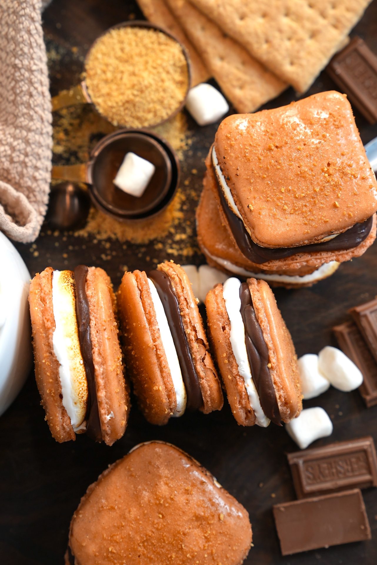 A close-up of several s’mores with toasted marshmallow and melted chocolate sandwiched between graham crackers, surrounded by loose marshmallows, chocolate pieces, graham crackers, and graham cracker crumbs.