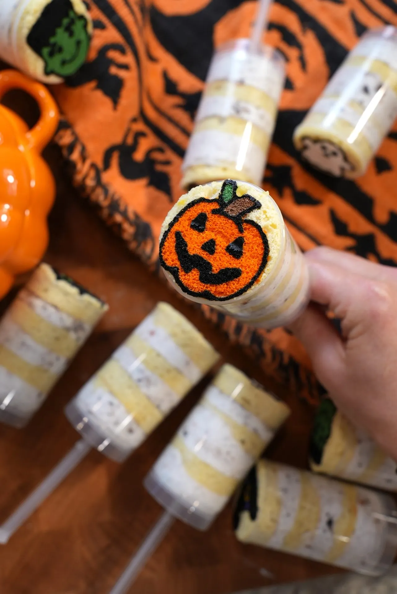 Hand holding a push pop cake with a pumpkin face on top; several similar Halloween-themed cake pops and orange pumpkin decor are in the background on a festive cloth.