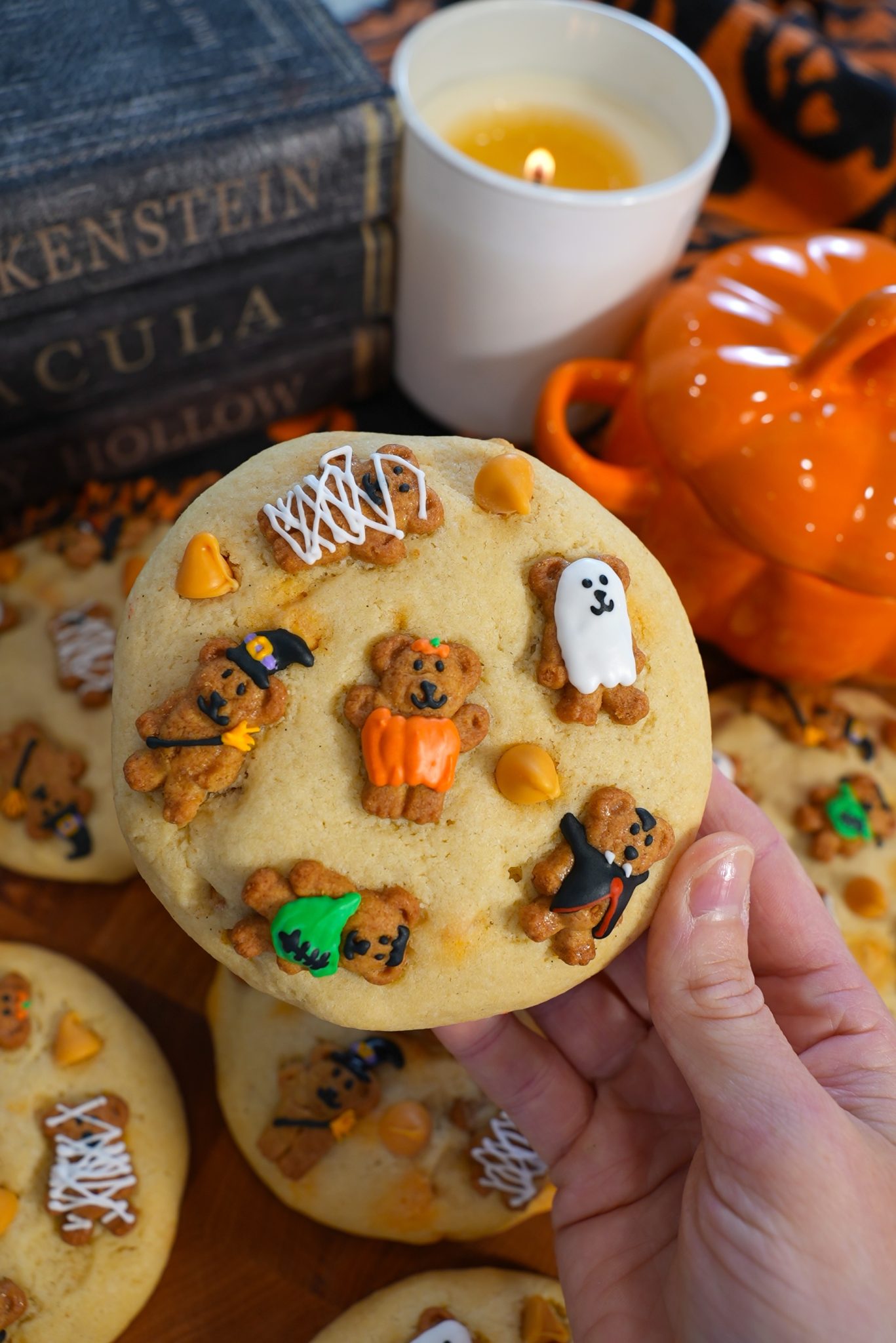 A hand holds a Halloween-themed cookie topped with bear-shaped cookies decorated as a pumpkin, ghost, witch, and vampire. More decorated cookies, old books, a lit candle, and an orange pumpkin dish are in the background.